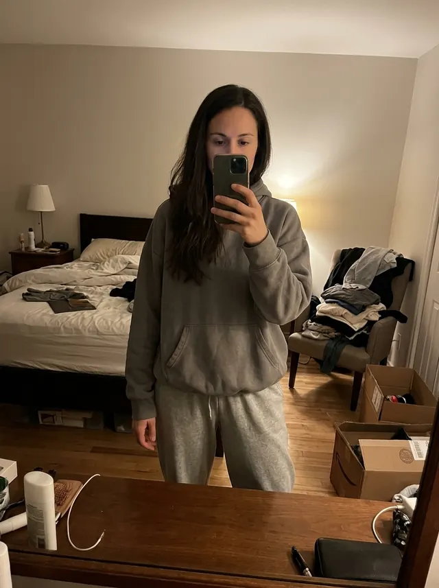 The same woman taking a mirror selfie in a cluttered bedroom, phone in front of her face, wearing an oversized grey hoodie and sweatpants
