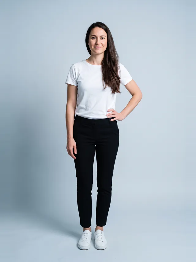 A woman standing straight and facing the camera in a fitted white t-shirt and slim black trousers, full length, against a plain pale backdrop